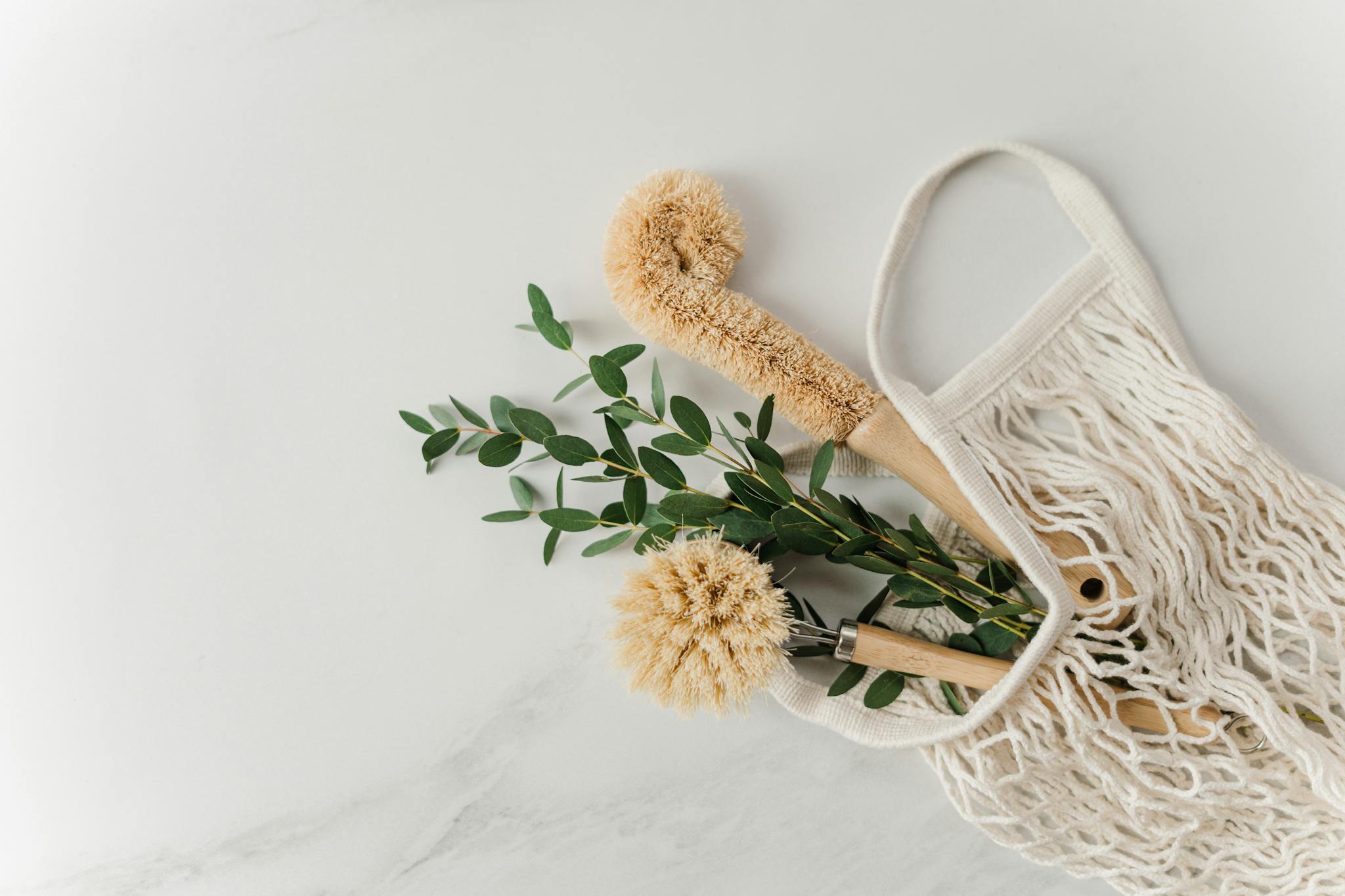 Eco-friendly cleaning brushes and eucalyptus branches in a reusable net bag on white surface.